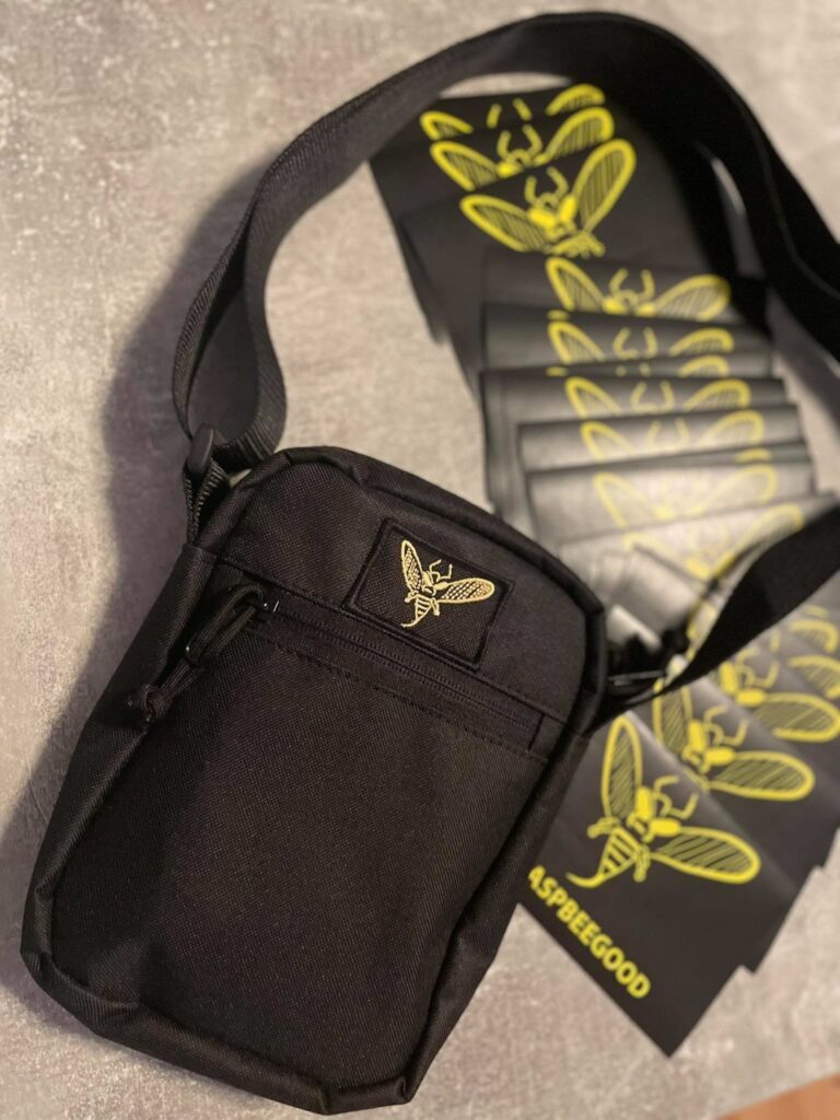 Mała czarna torba na ramię (listonoszka) ze złotym haftem i naklejkami reklamowymi. Kompleksowa produkcja gadżetów i toreb z logo dla firm (Custom Drop).