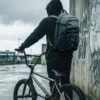 Plecak-droogBMX-Spot-Hunter-Rider-Street Rider w czarnej bluzie z kapturem stoi tyłem, mając na plecach oliwkowo-czarny plecak droogBMX Spot Hunter. Trzyma kierownicę roweru BMX na tle betonowej ściany z graffiti w pochmurny dzień.