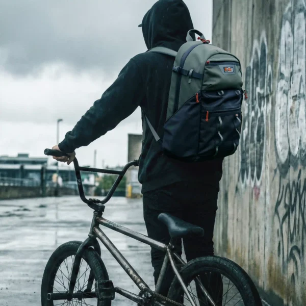 Plecak-droogBMX-Spot-Hunter-Rider-Street Rider w czarnej bluzie z kapturem stoi tyłem, mając na plecach oliwkowo-czarny plecak droogBMX Spot Hunter. Trzyma kierownicę roweru BMX na tle betonowej ściany z graffiti w pochmurny dzień.